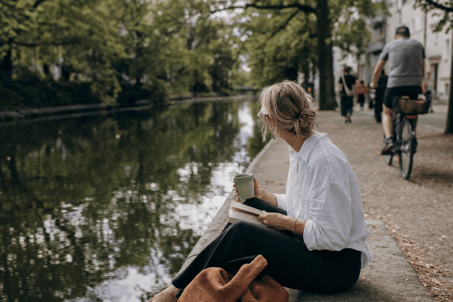 Eine Frau sitzt mit einem Buch und einem Becher am grünen Ufer des Landwehrkanals.