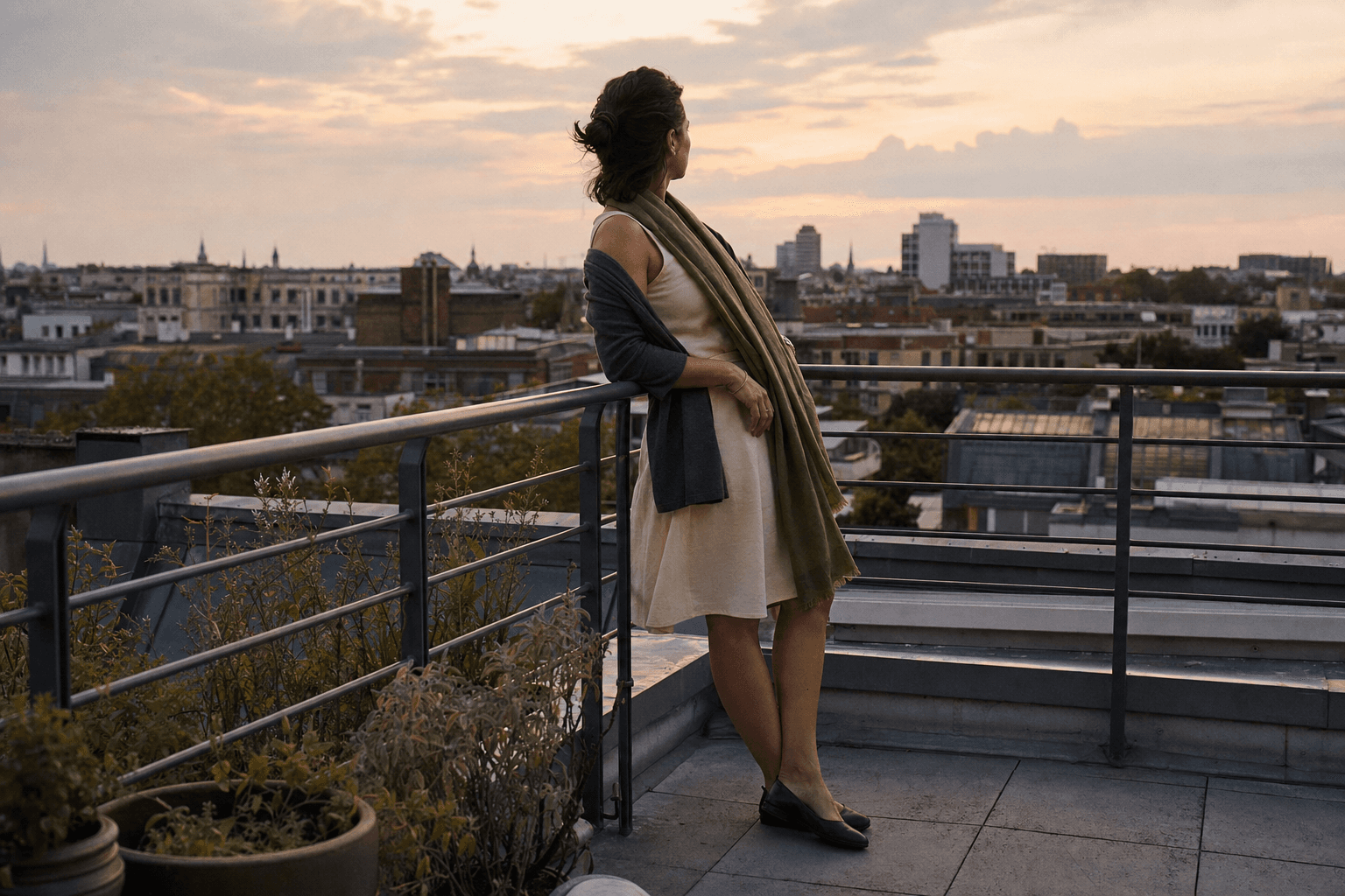 Eine Frau trägt auf einer Berliner Dachterrasse ein helles Kleid, eine dunkle Strickjacke und einen olivfarbenen Schal.